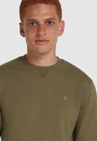 Olijfgroene sweatshirt met een ronde hals, gemaakt van zacht materiaal. Is voorzien van een klein oranje logo op de linkerkant van de borst en heeft een textuurdetail in de stiksels.