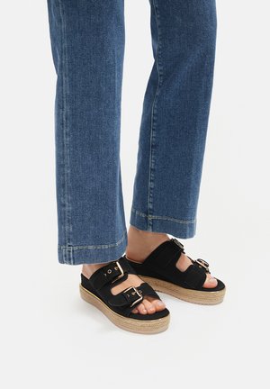 Czarne sandały na klinie z dwoma paskami na klamry i teksturowaną, plecioną podeszwą espadrylową. Noszone z niebieskimi szerokimi dżinsami.