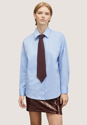 Camicia azzurra chiara con colletto a punta, abbinata a una cravatta marrone scuro. Sotto, una mini gonna marrone lucida con paillettes.