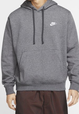 Mężczyzna w szarej bluzie z kapturem Nike z przednią kieszenią i sznurkami, w połączeniu z ciemnymi spodniami w kratę, pokazany od szyi do bioder.