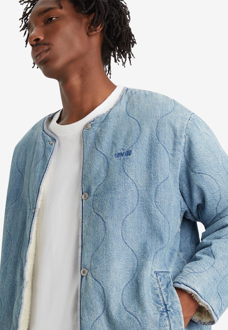 Chaqueta de denim en azul claro con diseño de olas, botones a presión y forro de felpa. Llevada sobre una camiseta blanca, con detalle de logo bordado.