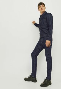 Chaqueta de mezclilla oscura con cierres de botón, combinada con jeans oscuros. Llevada con zapatos negros con cordones. Tela suave, diseño estructurado.