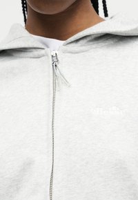 Makro záber osoby v svetlosivej mikine na zips s dvojitým zipsom a bielym tričkom pod ňou.