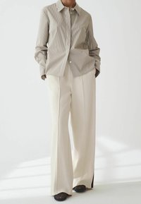 Personne portant une chemise beige à fines rayures rentrée dans un pantalon crème taille haute à jambes larges, avec des chaussures noires, se tenant devant un fond blanc.