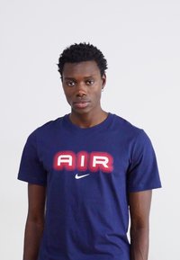 Κοντομάνικο μπλουζάκι από βαμβάκι ναυτικού μπλε, με έντονη κόκκινη και λευκή γραφική απεικόνιση "AIR", στρογγυλή λαιμόκοψη και λευκό λογότυπο Nike στη αριστερή πλευρά.