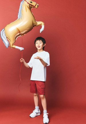 Un enfant en chemise blanche et short rouge tient un ballon gonflable doré en forme de cheval sur un fond rouge. Le ballon a une finition brillante.