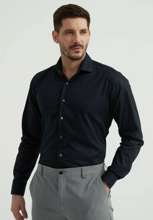 Chemise à manches longues bleu marine foncé avec un design à boutons, dotée de boutons bleus, coupe ajustée et tissu doux. Associée à un pantalon gris clair.