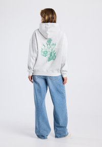 Gray hoodie with a green botanical print and text on the back. Paired with loose blue denim jeans. Cotton fabric with a relaxed fit.
