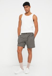 Canotta bianca abbinata a pantaloni corti neri con motivi circolari. Il modello indossa sneakers bianche e calzini chiari, in piedi dritto.