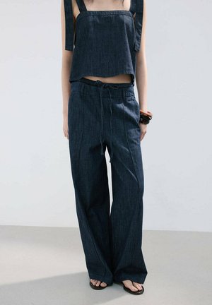 Person wearing dark blue loose-fit pants with drawstring waist, matching cropped top with wide shoulder straps, sandals, and bracelets.