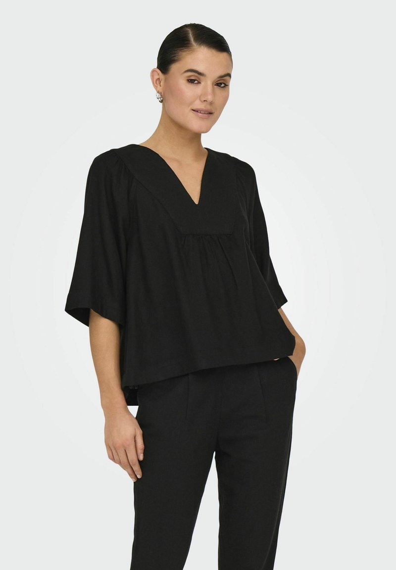 Top nero con scollatura a V, maniche corte e larghe. Presenta dettagli arricciati, vestibilità ampia, tessuto leggero e un drappeggio sottile. Abbinato a pantaloni neri.