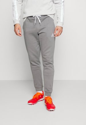 Osoba nosząca szare spodnie dresowe Adidas ze sznurkiem i czerwone korki piłkarskie z pomarańczowymi sznurowadłami, stojąca na jednolitym tle.