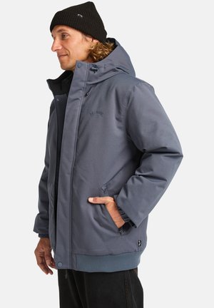 Giacca isolante grigia con chiusura a zip, grande cappuccio e polsini a costine. Include tasche laterali e una toppa con logo sul petto.