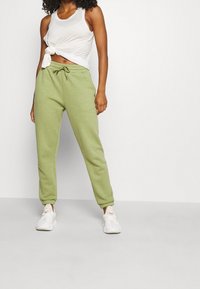 Světlé zelené tepláky s elastickými nohavicemi a pasem, doplněné uzlovým bílým tílkem. Bílé tenisky s texturovaným svrškem.