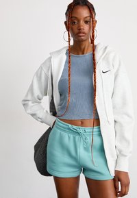 Ung kvinne med lange oransje fletter iført hoops-øredobber, grå hettegenser med glidelås, blå crop top og turkise shortser, som bærer en svart bag.