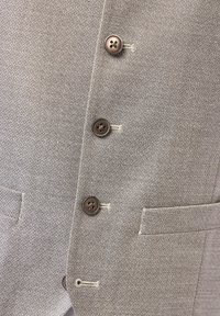 Hellgrau strukturierter Blazer mit fünf braunen Knöpfen, offenen Taschen und subtilen geometrischen Mustern. Taillierte Passform mit sauberen Nähdetails.