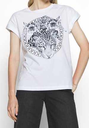 Femme portant un t-shirt blanc avec un visage de léopard noir entouré de fleurs dans un motif géométrique circulaire, associé à un pantalon foncé.