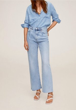 Kobieta ubrana w jasnoniebieskie denimowe spodnie z wysokim stanem i szerokimi nogawkami, dopasowaną jasnoniebieską koszulę zapinaną na guziki, z podwiniętymi rękawami, oraz białe sandały na paski.