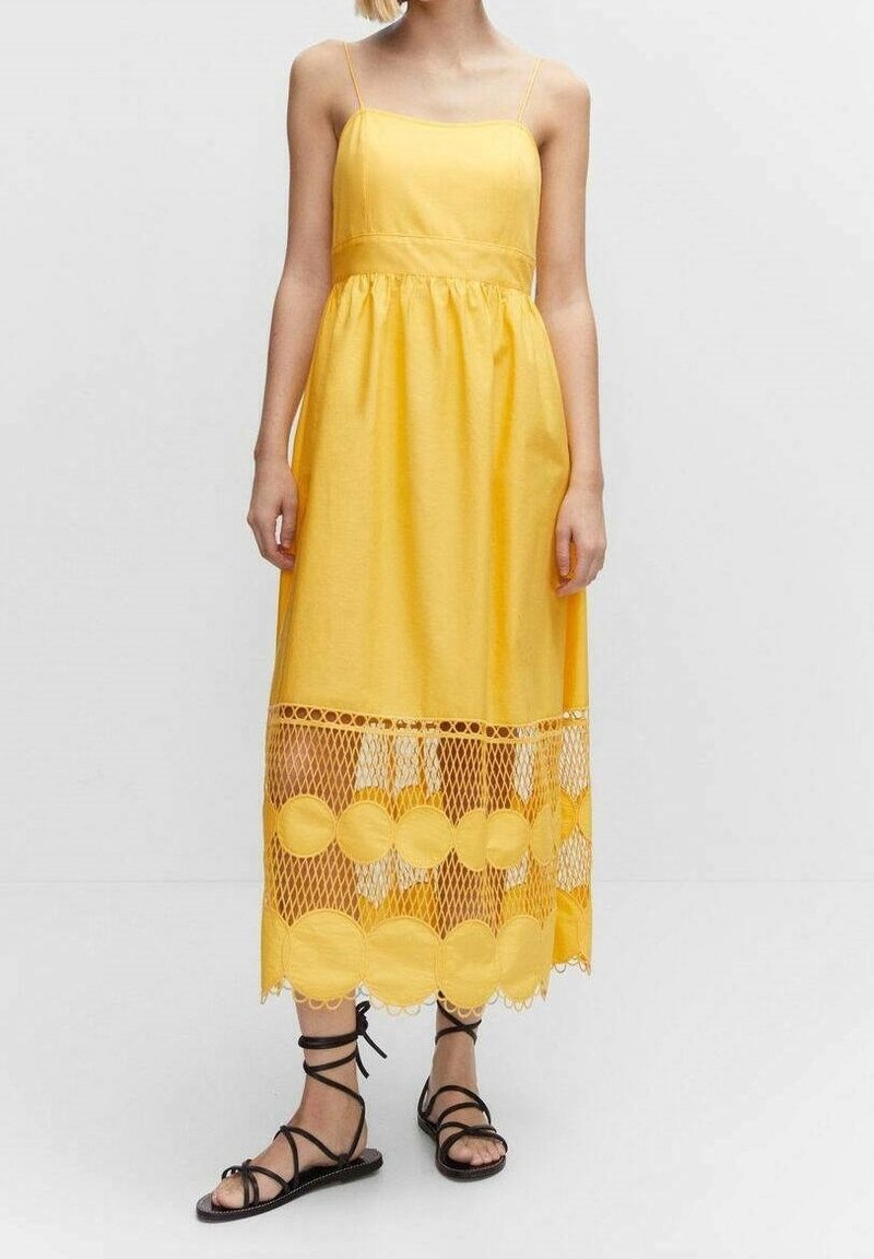 Femme portant une robe midi jaune sans manches avec de fines bretelles et un ourlet en dentelle résille, associée à des sandales plates noires à brides fines.