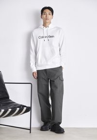 Witte hoodie met "Calvin Klein" logo, gecombineerd met donkergrijze cargobroek en zwarte sneakers. Het heeft een soepele stof met een casual pasvorm, inclusief zakken.