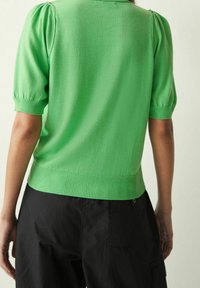 Groene gebreide trui met korte pofmouwen en ribbels aan de onderkant. Zachte textuur, getailleerd ontwerp, gedragen met zwarte broeken, vanuit de achterkant gezien.