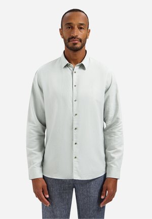 Mann steht, trägt ein hellgraues, langärmeliges Hemd mit Knöpfen und dunkelgraue Hose, schaut nach vorne vor weißem, einfarbigem Hintergrund.