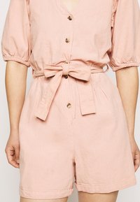 Ljust rosa jumpsuit med korta puffärmar, knäppning fram, avskuren midja med knytbälte och sidofickor. Mjuk tyg med subtil textur.