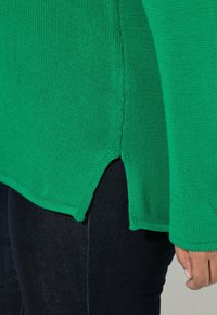 Maglione verde lavorato a maglia con maniche lunghe, caratterizzato da uno spacco laterale e una texture a coste. Abbinato a jeans denim blu scuro.