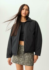 Giovane donna con lunghi capelli scuri, che indossa una giacca nera con zip, un top corto nero, una minigonna leopardata e orecchini a cerchio, posando contro uno sfondo bianco.