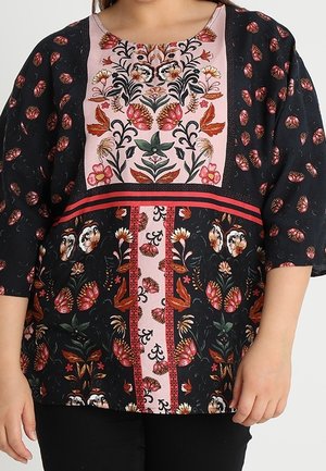 Kvinde iført en sort bluse med lyserøde paneler og farverigt blomsterbroderi med røde, hvide og grønne blomster- og bladmotiver.