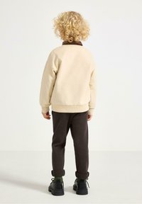 Beżowa bluza z brązowym kołnierzem, ciemnoszare spodnie i czarne buty. Widoczne kręcone włosy. Skoncentrowanie na luźnym kroju i miękkich fakturach.