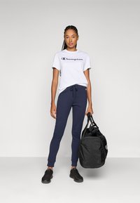 Witte T-shirt met "Champion" logo, marineblauwe joggingsbroek met koord, zwarte sneakers en een zwarte sporttas. Glad materiaal, sportieve pasvorm.