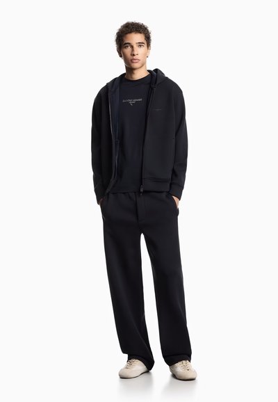 Sudadera con cremallera negra sobre una camiseta negra, combinada con pantalones negros holgados y zapatos de color claro. Tejido suave con mínima marca.