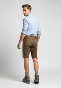 Pantaloni corti in suede marrone con vita regolabile e dettagli laterali, abbinati a una camicia azzurro chiaro con bottoni e stivali da trekking marroni.