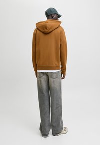 Férfi barna kapucnis pulóverben, szürke bő szárú farmernadrágban, fehér tornacipőben és zöld sapkában, háttal áll egy egyszerű háttér előtt.