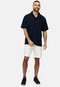 Camisa de manga corta texturizada en azul oscuro con cuello, combinada con pantalones cortos blancos arremangados y zapatillas negras. Detalles de botones en la camisa.