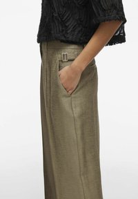 Pantalones de tiro alto beige con una textura suave, bolsillos laterales y un acento de hebilla plateada en la cintura. Blusa negra arrugada parcialmente visible.