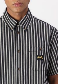 Striped short-sleeve shirt in black and white cotton. Features a button-down collar, brown buttons, and a left chest pocket with logo.
