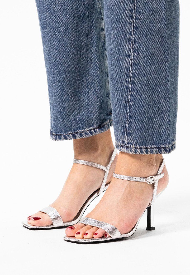 Zilveren metallic high-heeled sandalen met een minimalistisch ontwerp, voorzien van dunne banden en een vierkante neus. Slanke enkelbanden hebben een decoratieve gesp.