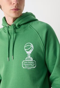 Πράσινη φούτερ με κεραμιδί σχέδιο τροπαίου και μπάσκετ. Διαθέτει κορδόνια με μεταλλικές άκρες και εκτυπωμένο κείμε�νο: "Πρωτάθλημα Lower Manhattan 11 Μαρτίου 1990."