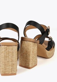 Sandalias de plataforma de cuero negro con correas anchas y un tacón de bloque de yute trenzado. El tacón tiene una textura natural y una base marrón resistente.