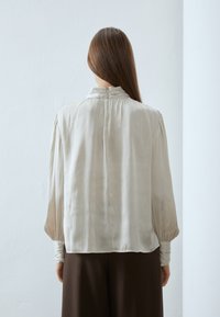 Lys beige bluse med en myk, silkeaktig tekstur. Har høyt krage og rynkede detaljer ved skuldrene og mansjettene. Passer godt sammen med brune bukser.