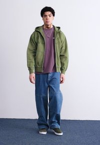 Green zip-up hoodie, maroon shirt underneath, oversized blue denim pants, and green shoes. Simple design without patterns or notable accents.