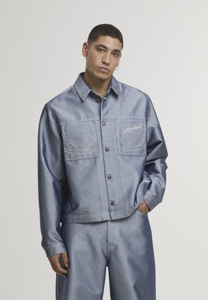 Noor mees kannab helehalli üle suuruses teksariidest jopet ja sobivaid laiade säärega teksapükse, seisab ühel käel taskus tavalise taustaga tausta ees.