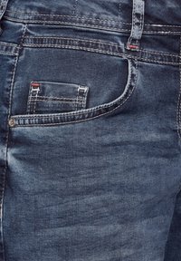 Jeans de mezclilla oscura con un bolsillo trasero con costura visible, un pequeño bolsillo arriba y un acento de botón metálico. Textura suave.