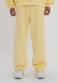 Pantalones de chándal amarillos de corte holgado, fabricados con un tejido suave. Presentan puños elásticos y un pequeño logo blanco en el costado. Combinados con zapatillas blancas.