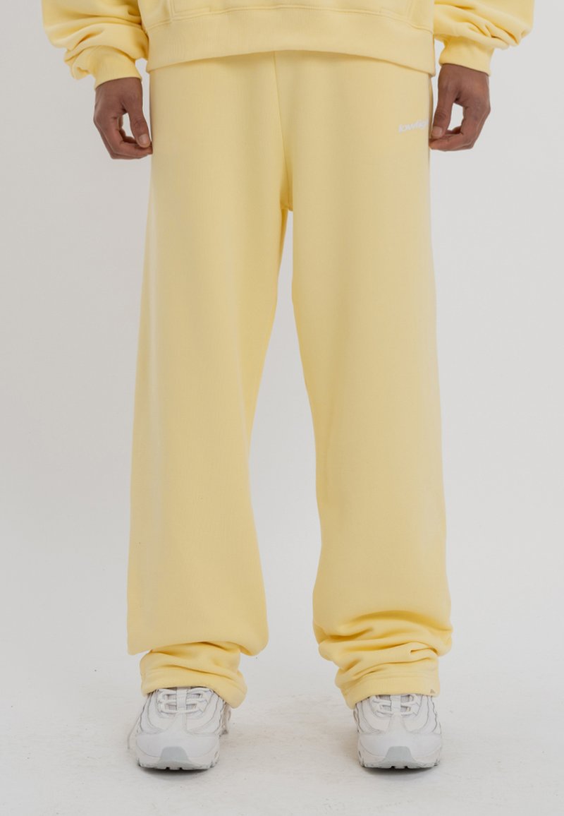 Pantalones de chándal amarillos de corte holgado, fabricados con un tejido suave. Presentan puños elásticos y un pequeño logo blanco en el costado. Combinados con zapatillas blancas.