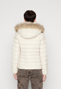 Vaaleanvärinen toppatakki, jossa on tekoturkiksinen huppu. Ominaisuuksiin kuuluu tikattu muotoilu ja resorit hihansuuissa. Takakuvassa näkyy vartalonmyötäinen siluetti.