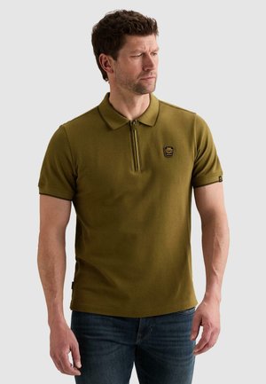 Homme portant un polo à manches courtes vert olive avec une fermeture éclair à l’avant et un patch logo, associé à un jean bleu foncé, regardant sur le côté.