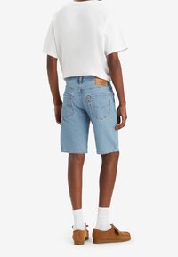 Pantalones cortos de mezclilla azul claro con corte recto, que cuentan con bolsillos traseros y un parche de logo de cuero. Combinados con una camiseta blanca y zapatos marrones.